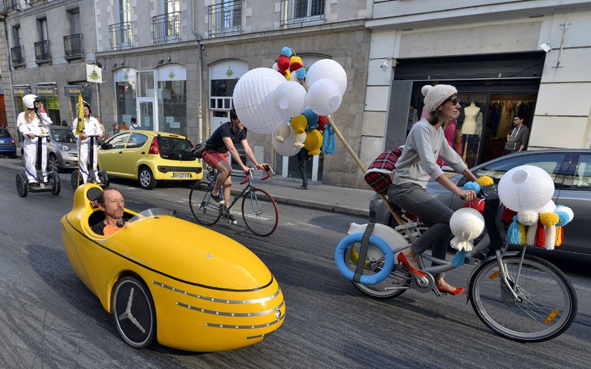 Biciklijada u Velo-gradu u Nantu - žena vozi bickl ukrašen balonima pored čovjeka u mini trkaćem automobilu